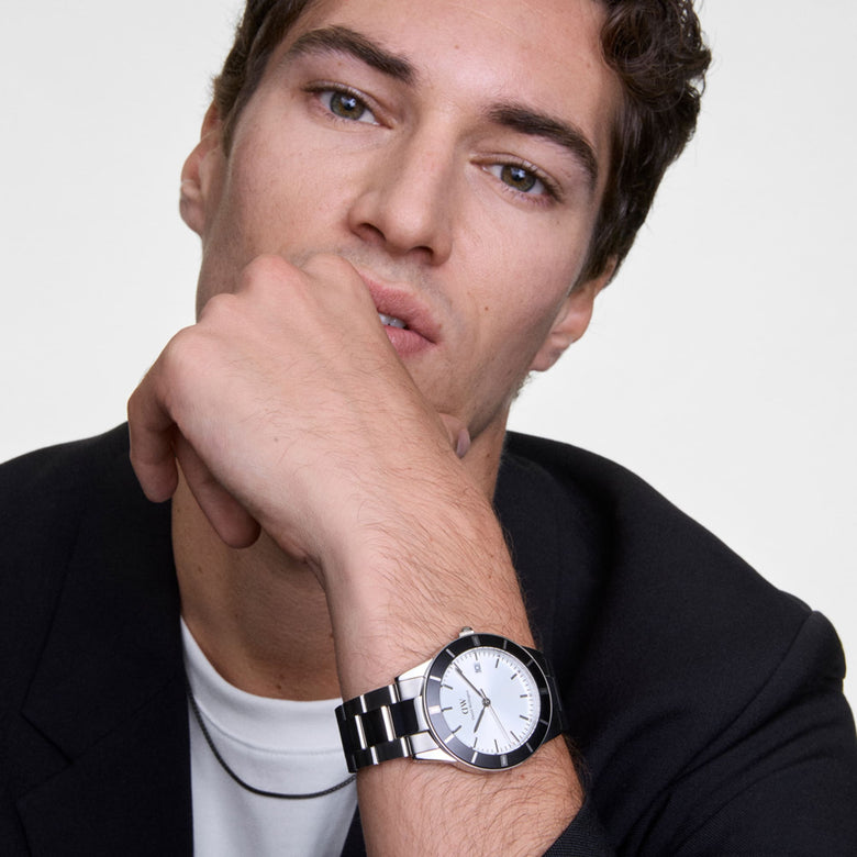 A man wearing the Daniel Wellington Iconic Paradigma Link Black Bezel watch with a stainless steel bracelet and bold black bezel, styled with a black blazer and white T-shirt for a smart-casual look.