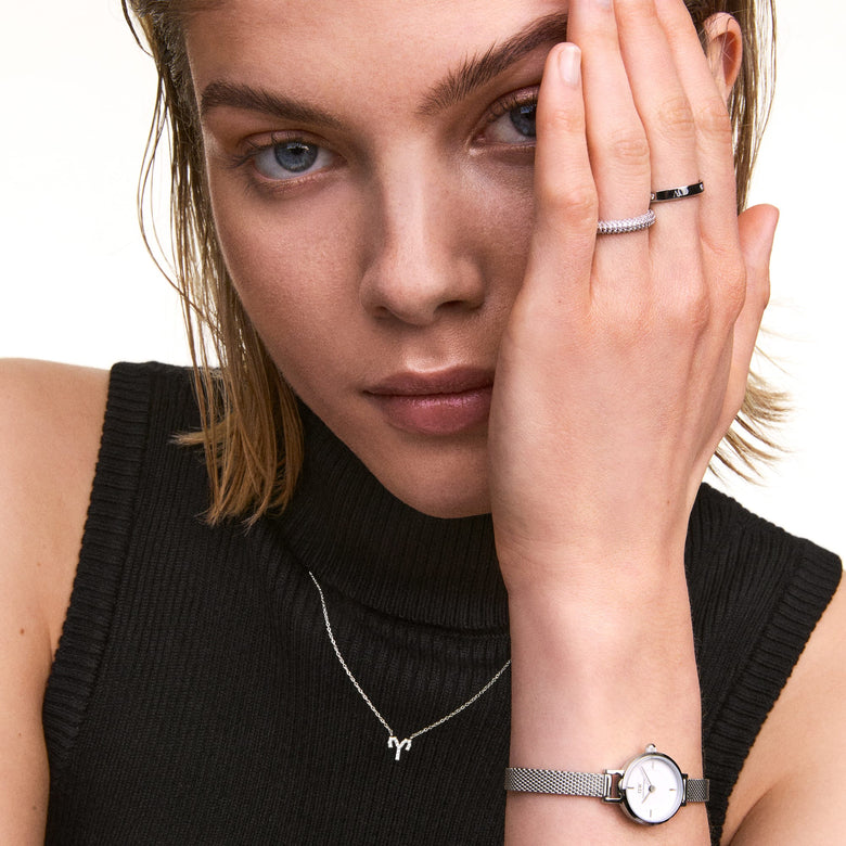 A woman wearing the Crystal Zodiac Chain Necklace Rose Gold, styled with a sleeveless black top, silver rings and a mesh strap watch, showcasing the delicate zodiac pendant as a subtle, modern accessory.