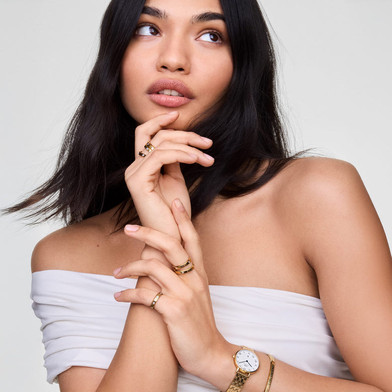 A woman wearing the Petite Roman Numerals Date 5-link Gold watch styled with gold rings and a gold bracelet, highlighting the watch’s slim gold 5-link bracelet, white dial, Roman numerals, and blue hands for an elegant, modern look.