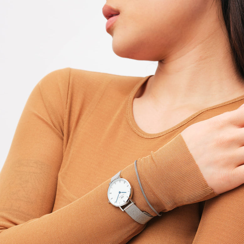 A woman wearing the Petite Roman Numerals Sterling watch with a silver mesh strap and white dial, styled with a long-sleeve tan top for a modern, everyday look.
