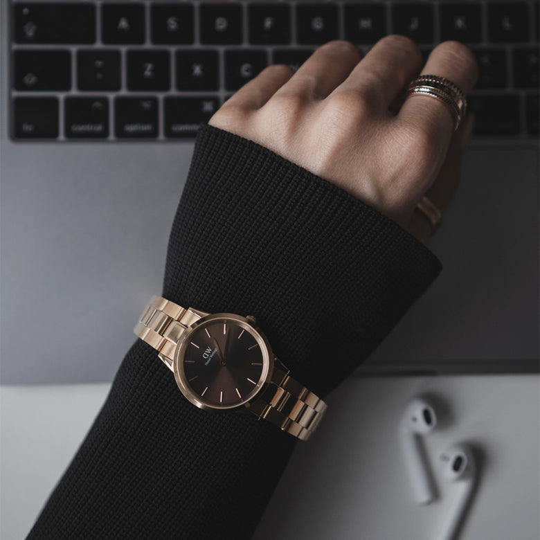 A person wearing the Daniel Wellington Iconic Link Amber watch with a rose gold bracelet and sunray brown dial, styled with a black jumper while working on a laptop.