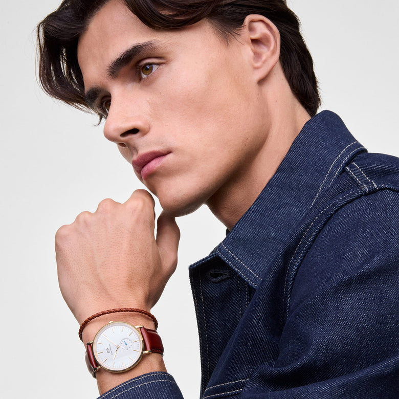 A man wearing the Classic Multi-Eye St Mawes Off-white Sunray Gold watch with a gold-plated round case, light beige sunray dial, slim gold markers, blue sub-second hand, and a stitched brown leather strap, styled with a denim jacket and braided bracelet.