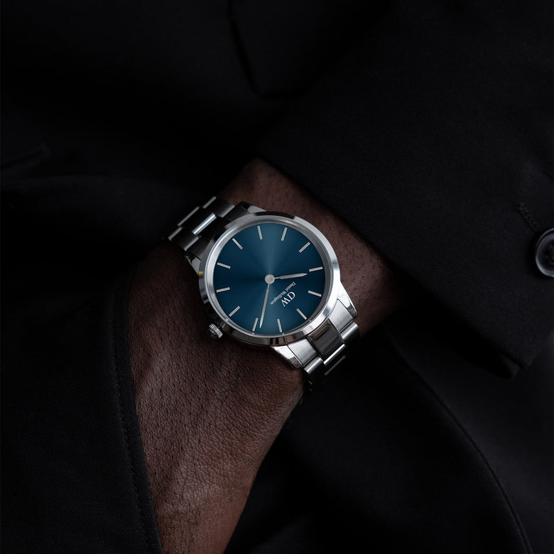 A Daniel Wellington Iconic Link Arctic Silver watch with a deep blue sunray dial and polished stainless steel bracelet, worn on the wrist with a formal black suit.