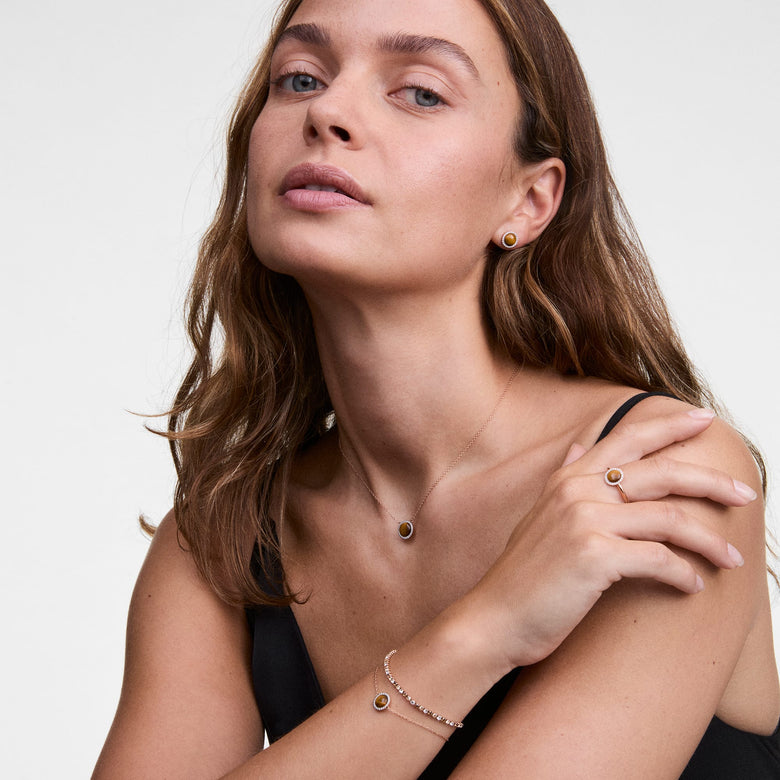 A woman wears Daniel Wellington Audrey Earrings Tiger Eye Gold, featuring round tiger eye stones framed by cubic zirconia, styled with matching gold jewellery for an elegant, coordinated look.