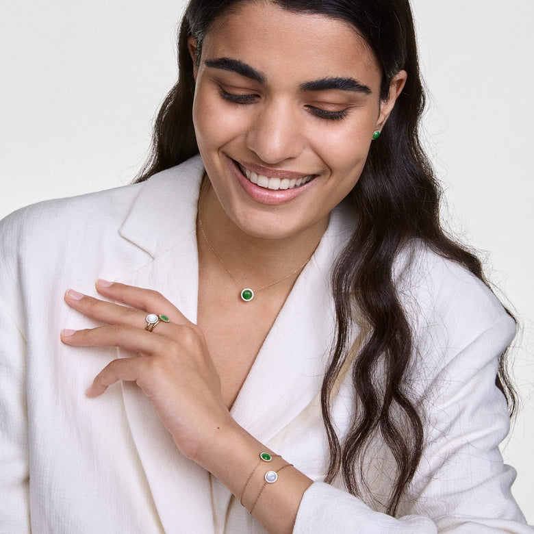 A woman wearing the Audrey Ring Jade Rose Gold, styled with a matching jade necklace and bracelet, showcases the ring’s green jade centrepiece and sparkling cubic zirconia halo against a white blazer for a modern, elegant look.