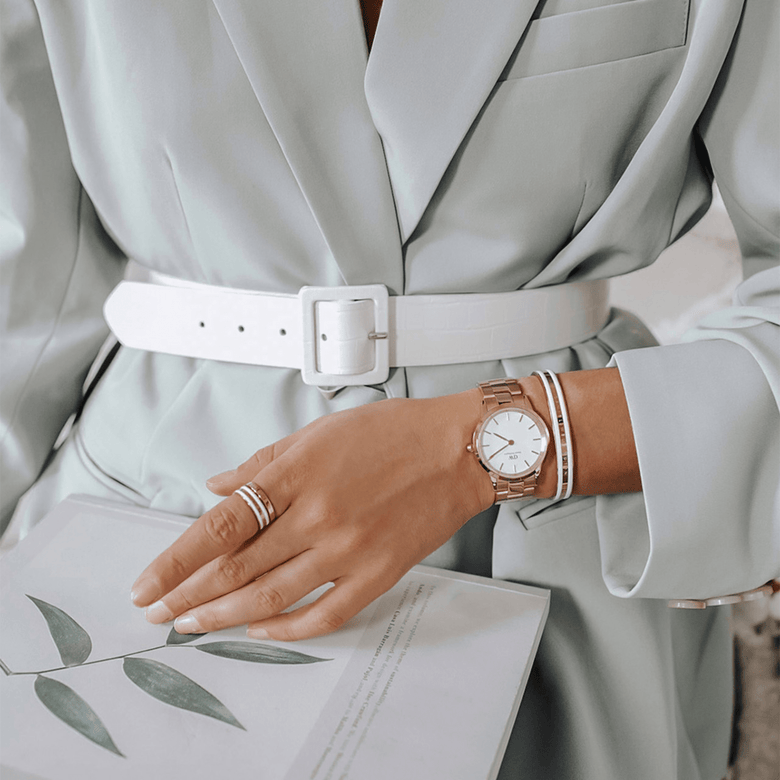 The Iconic Link Rose Gold watch with a polished rose gold three-link bracelet and minimalist eggshell white dial, styled on a wrist with a white bangle and pastel suit, creating a refined look for business or smart casual wear.