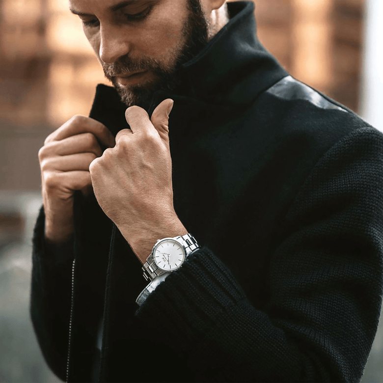 Iconic Link Silver watch by Daniel Wellington worn on the wrist with a black textured coat, showcasing its polished stainless steel bracelet and minimalist eggshell white dial in a stylish outdoor setting.