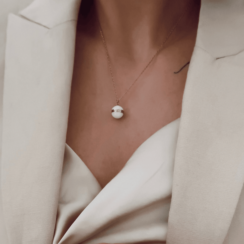 Aspiration Necklace Rose Gold Satin White worn on the collarbone, featuring a satin white ceramic pendant with a rose gold plated band, styled with an elegant cream blazer and top.