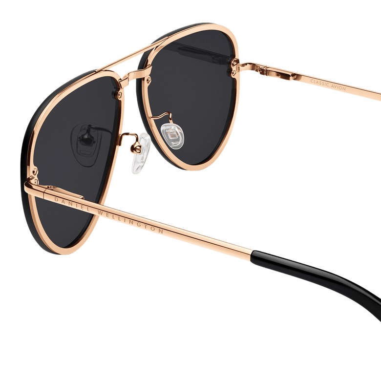 Close-up of Avion Steel Rose Gold sunglasses highlighting the rose gold plated stainless steel frame, black aviator lenses, adjustable silicone nose pads, and Daniel Wellington branding on the arm.
