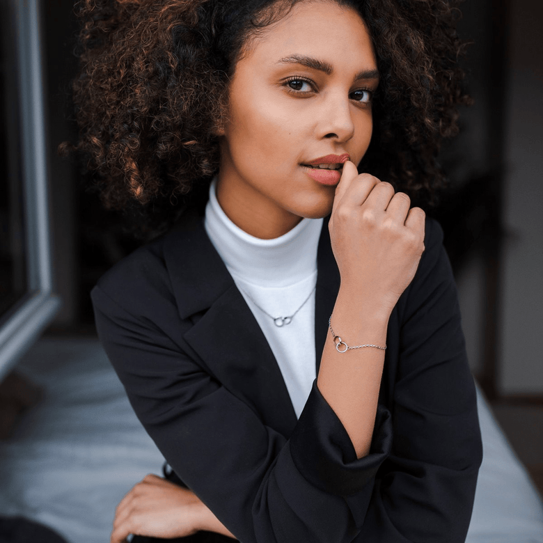 A woman wearing the Elan Unity Necklace Silver with two interlocked rings on a fine chain, styled over a white turtleneck and black blazer for a modern, elegant look.