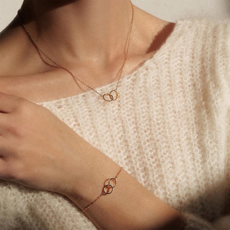 Woman wearing the Elan Unity Bracelet Rose Gold and matching necklace with interlocking rings, styled with a soft cream knit top for a classic and elegant look.